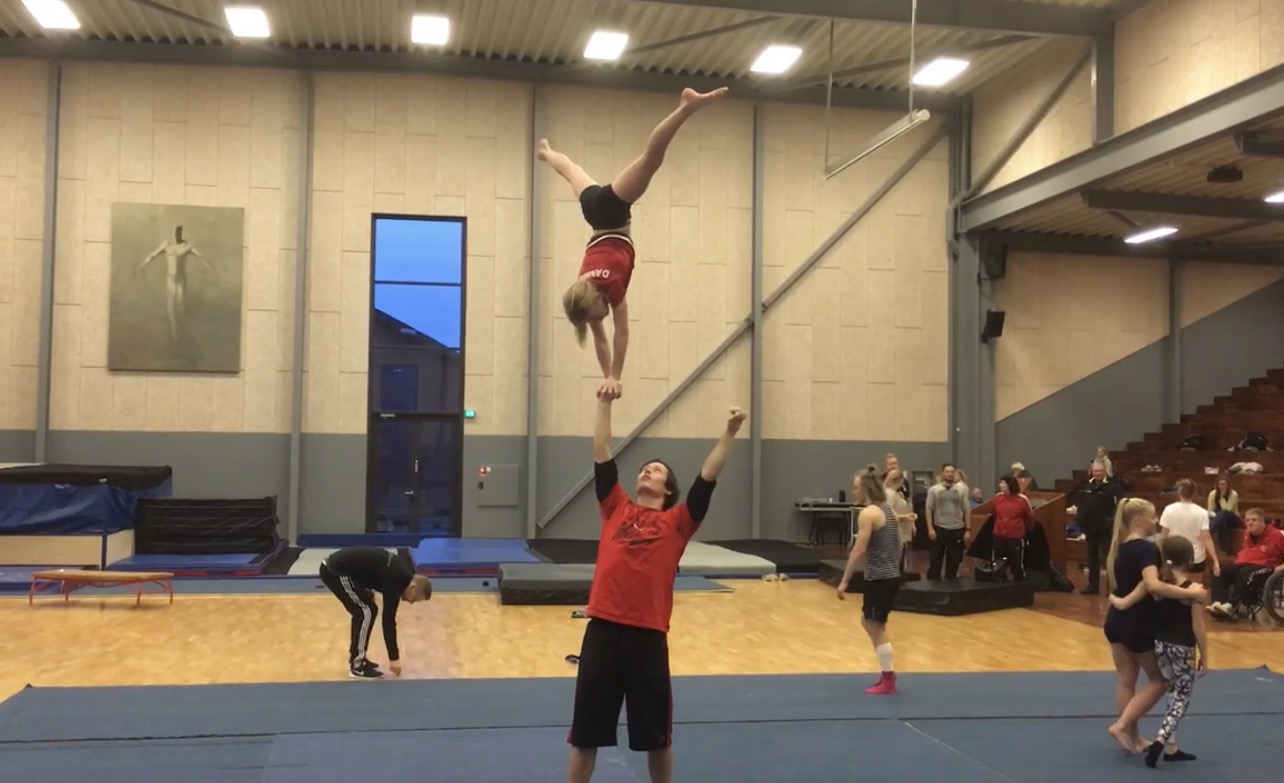 Didrik, wearing a red shirt, supporting an inverted partner during sports-acrobatics education in a gymnastics hall